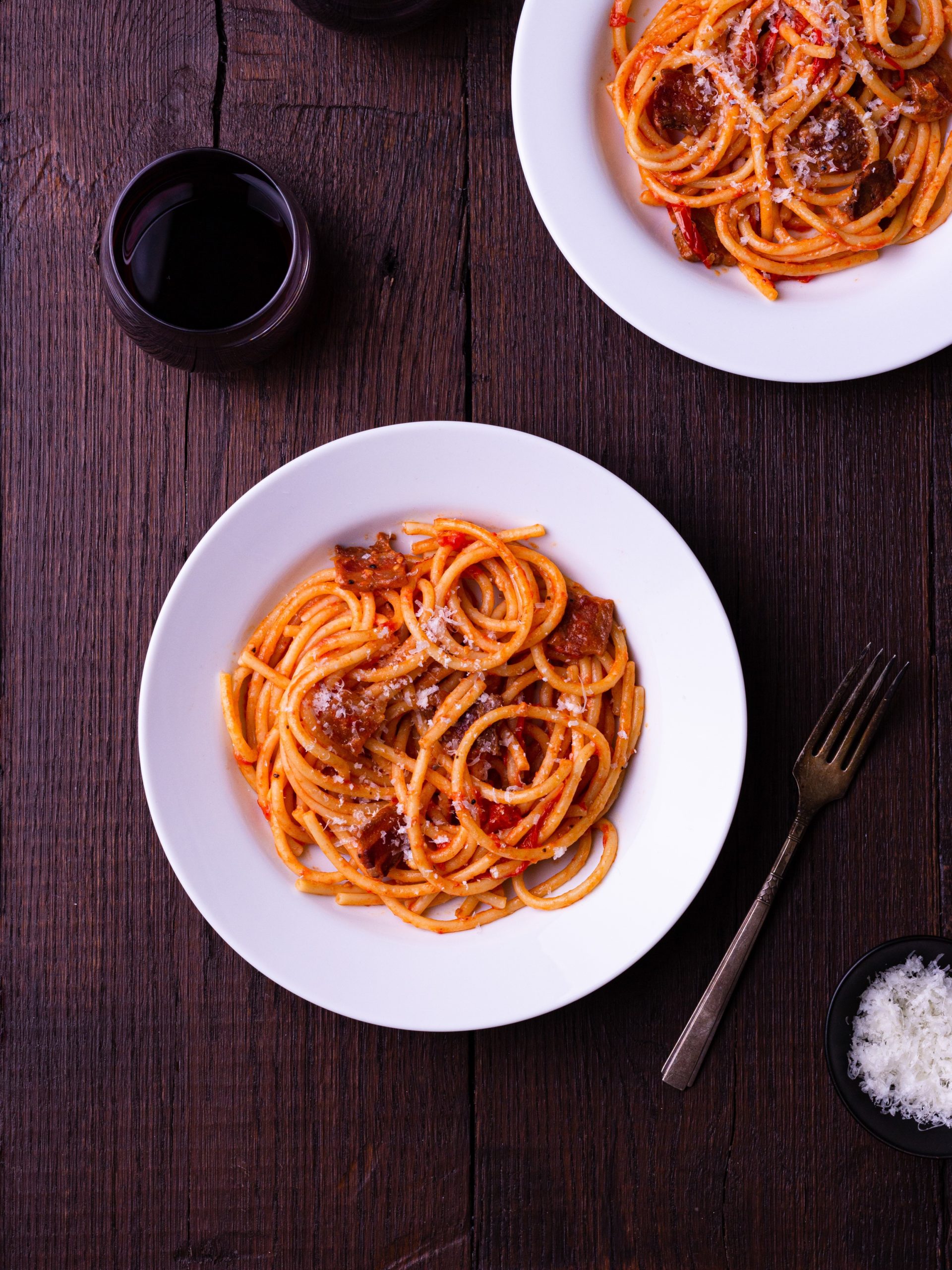 Two bowls of pasta with Amatriciana sauce topped with grated pecorino cheese, surrounded by glasses of red wine and a fork.