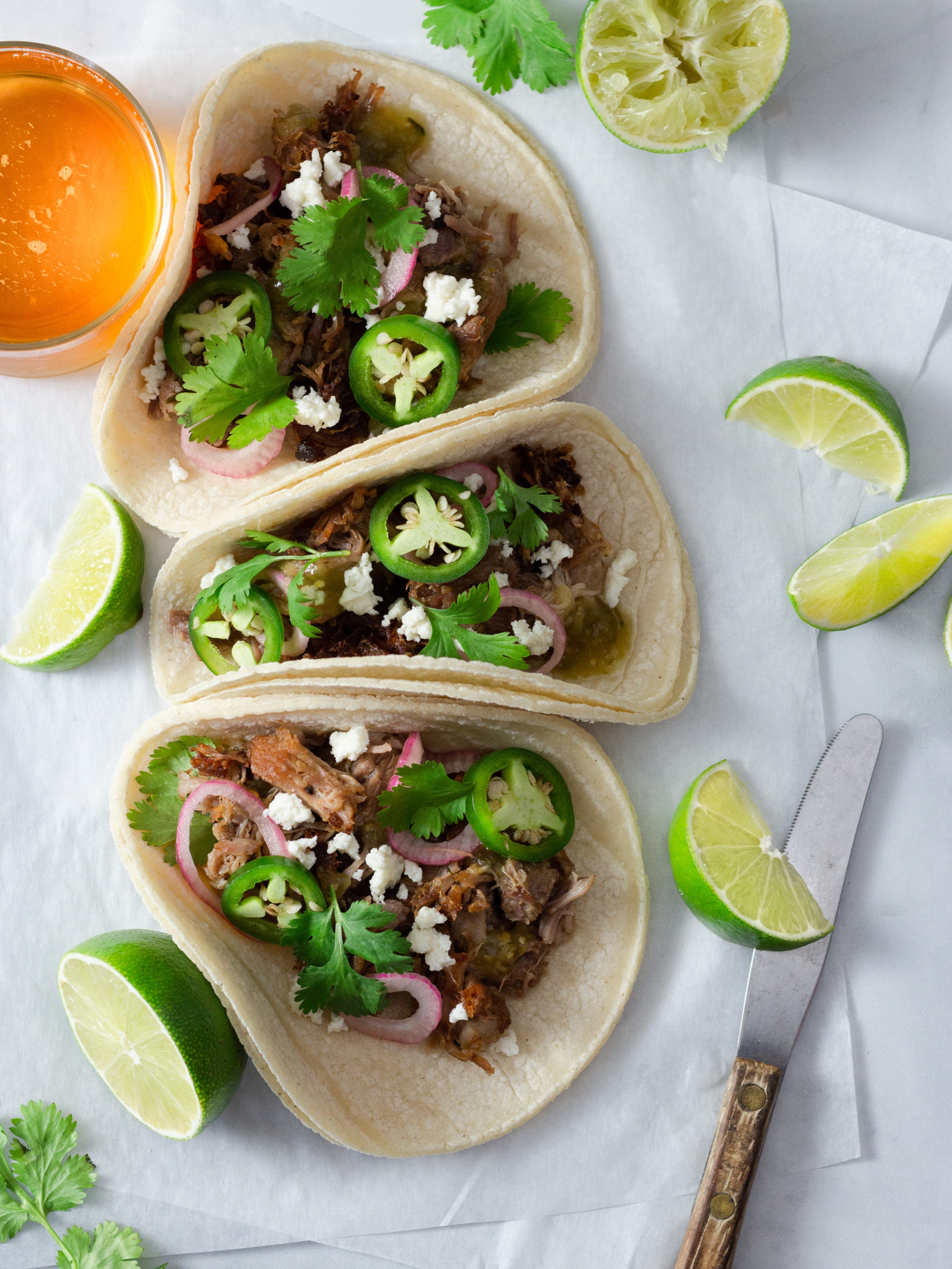 Overhead view of three carnitas tacos topped with red onions, jalapeños and cilantro, surrounded by limes and a beer.