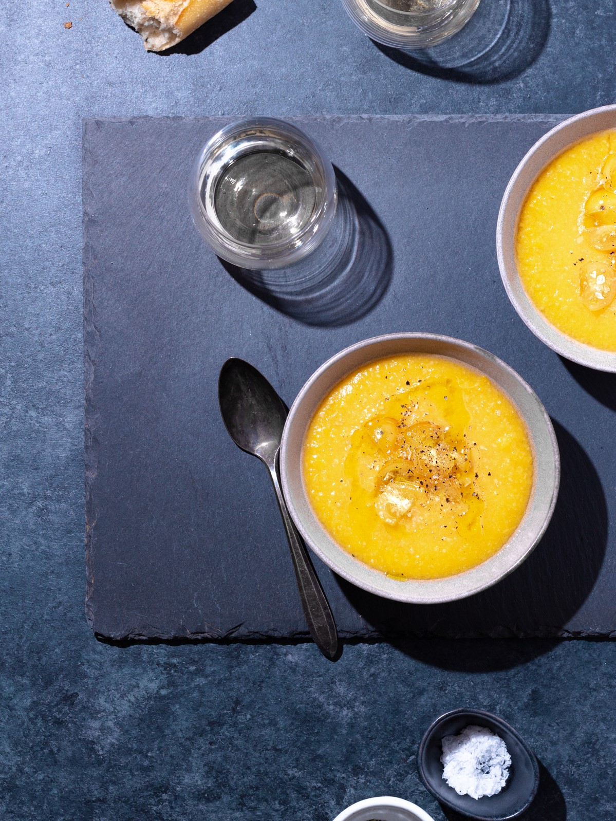 Overhead view of two bowls of yellow gazpacho and a grey-blue surface surrounded by glasses of white wine, crusty bread, a spoon and salt and pepper bowls.