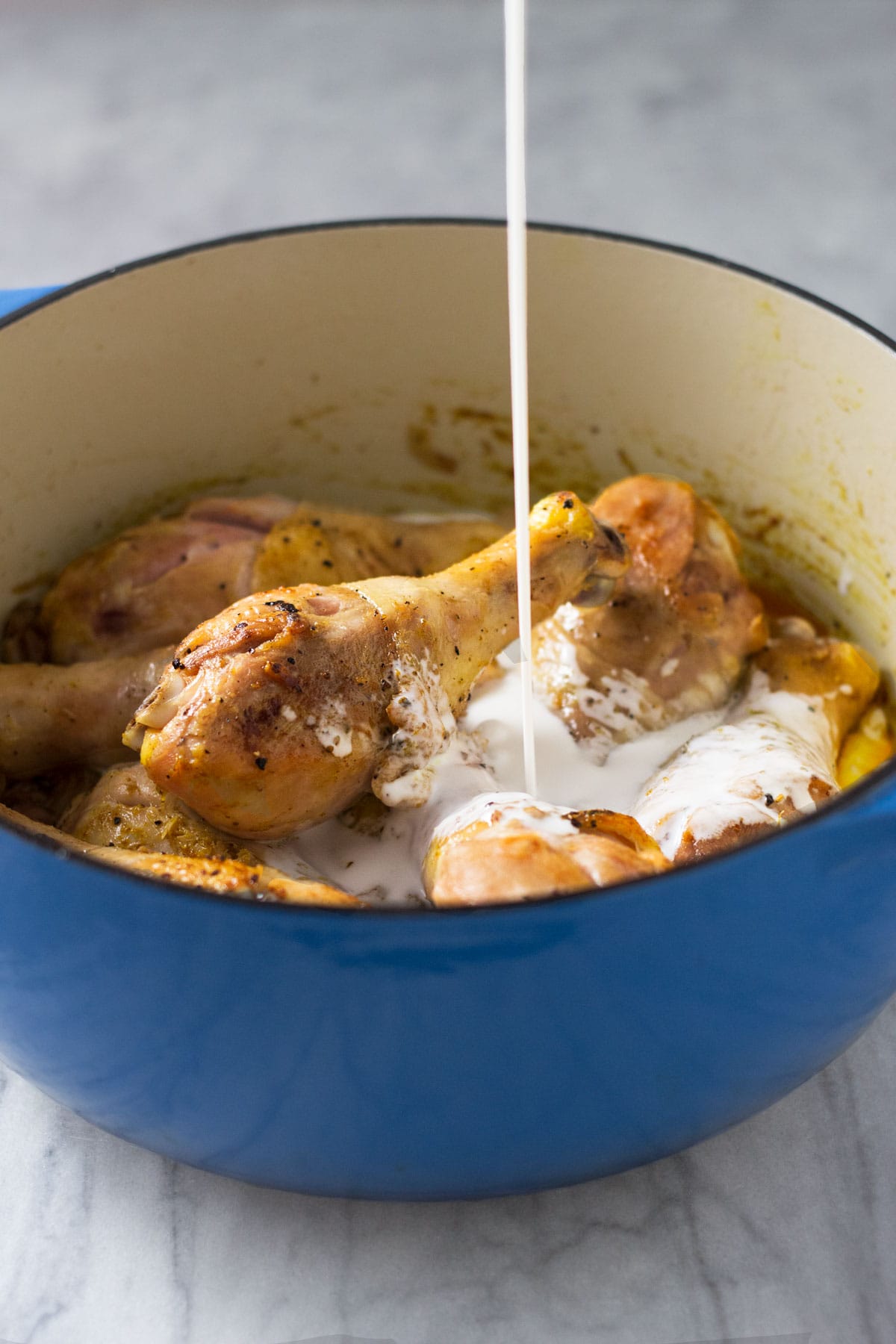 Straight on view of a pot of browned chicken legs with coconut milk being poured into pot.