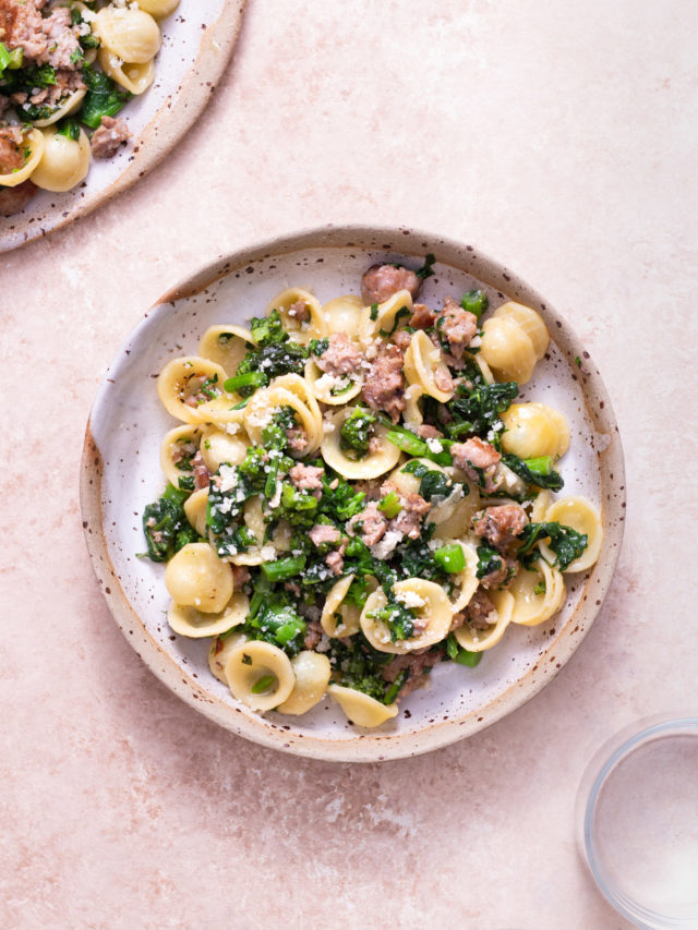 Overhead view of a bowl of orecchiette pasta with sausage, broccoli rabe and grated cheese on a textured light brown surface.