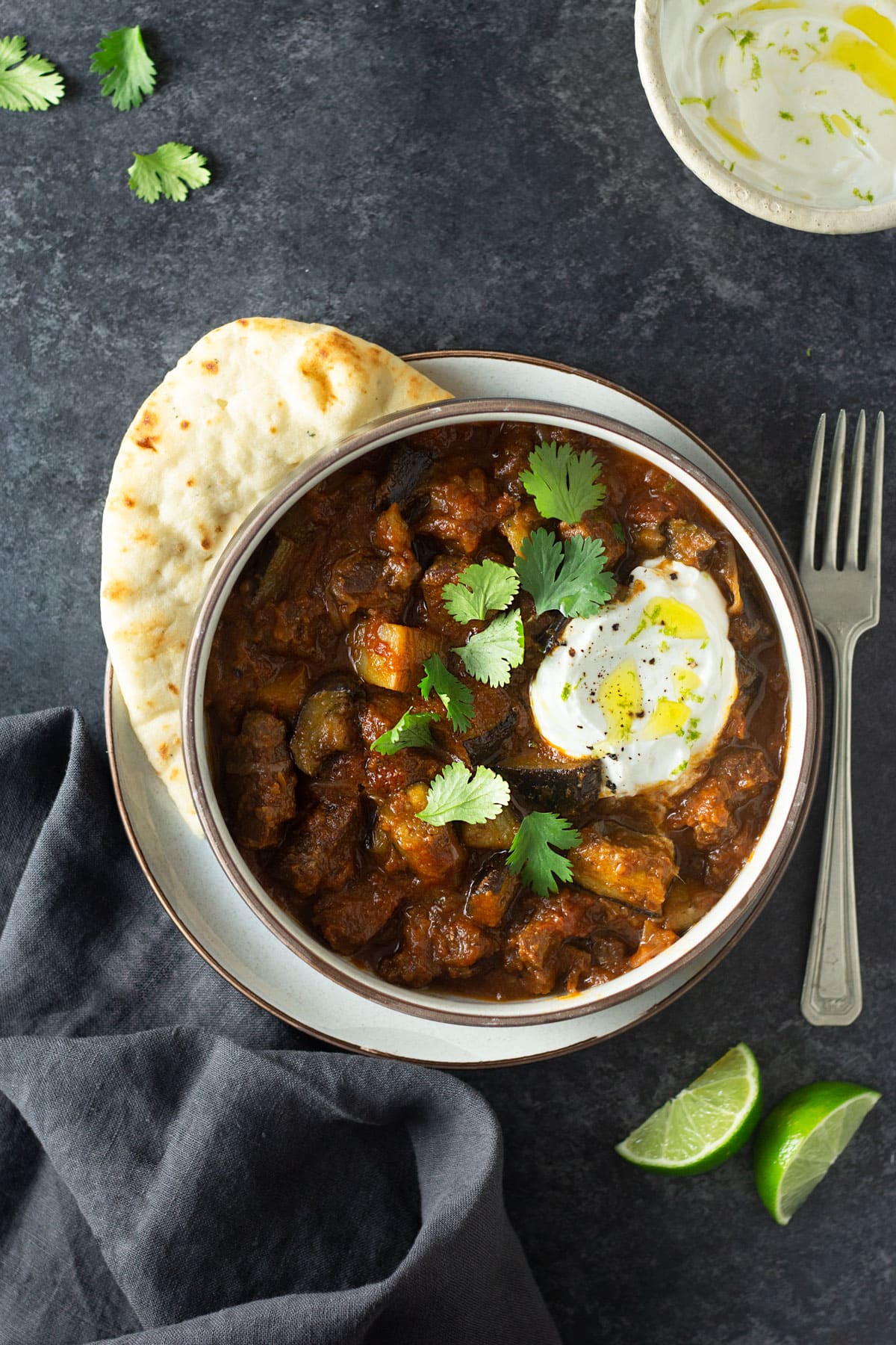 Overhead view of a bowl of Indian inspired lamb curry surrounded by yogurt, lime wedges and naan on a dark grey surface.