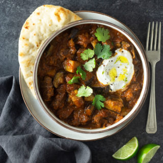 Overhead view of a bowl of Indian inspired lamb curry surrounded by yogurt, lime wedges and naan on a dark grey surface.