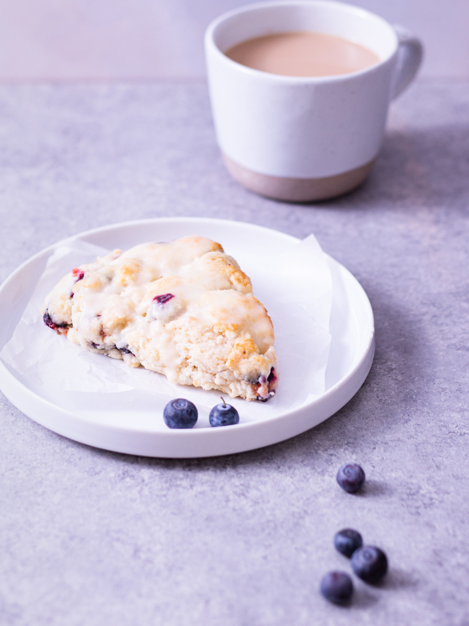 Straight on shot of a blueberry scone on a white plate next to a cup of coffee on a light grey surface.