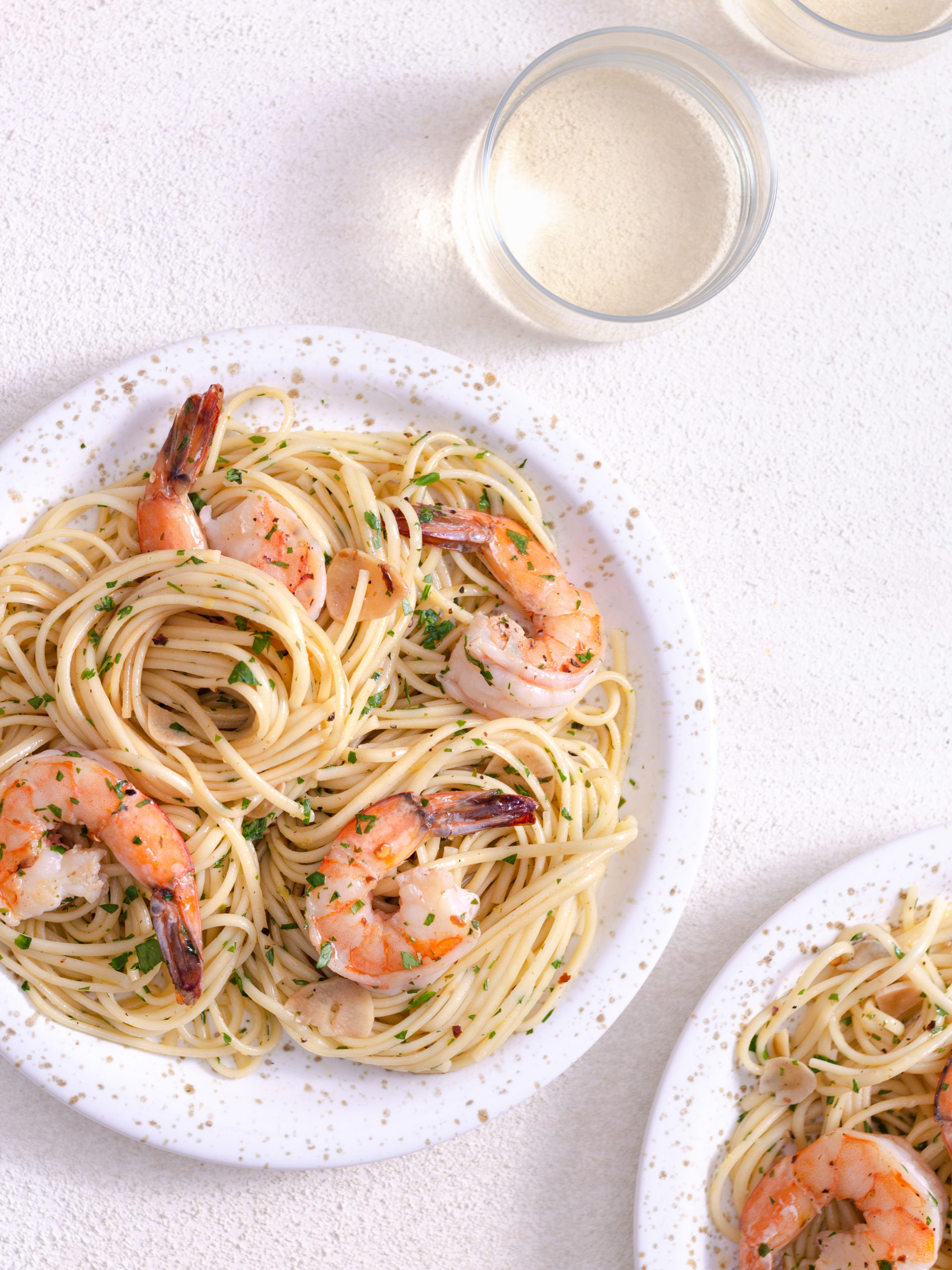 Overhead view of two plates of shrimp pasta on a light surface next to glasses of white wine.