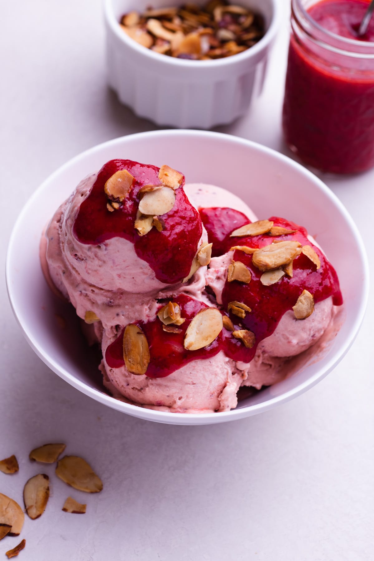Strawberry Balsamic Ice Cream in a bowl topped with strawberry sauce and toasted almonds with the toppings in containers on the side.