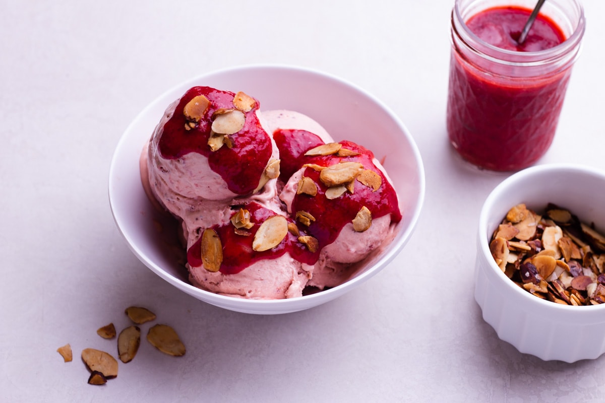 Strawberry Balsamic Ice Cream in a bowl topped with strawberry sauce and toasted almonds with the toppings in containers on the side.
