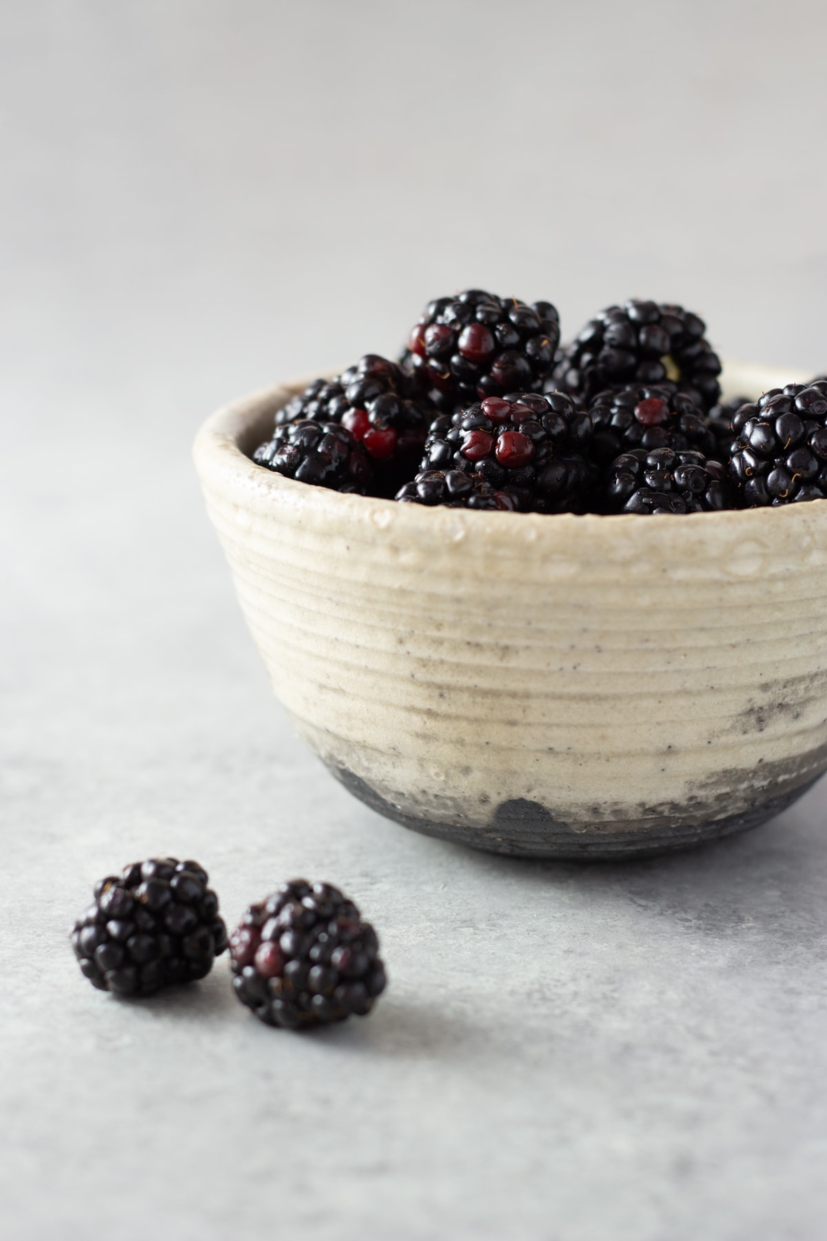 Straight on shot of a rustic bowl filled with fresh blackberries on a light grey surface.