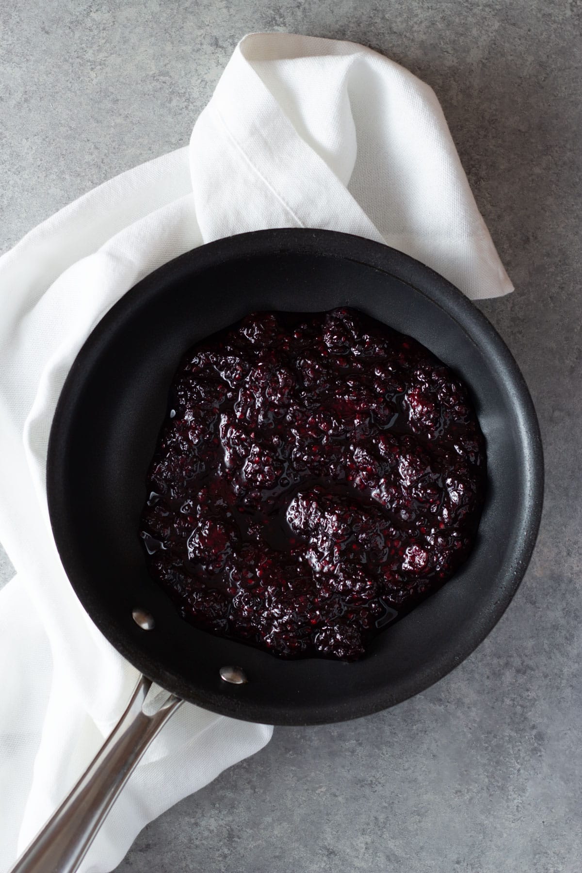 Overhead view of a pan with blackberry jam next to a dishtowel on a grey surface.