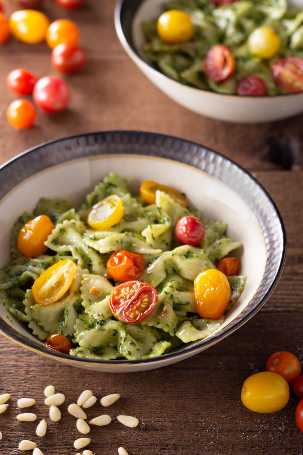 Angled view of a bowl of farfalle pasta with pesto sauce and roasted tomatoes on a wood surface.