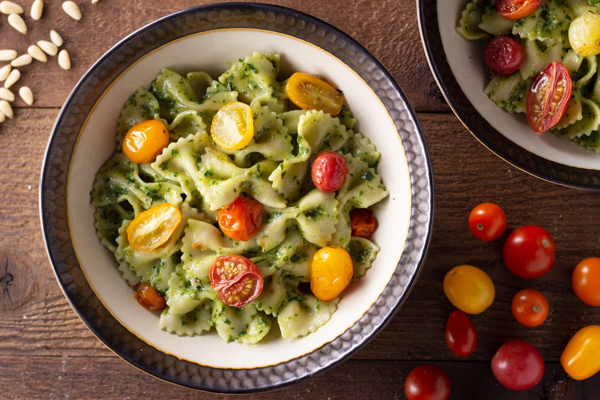Overhead view of two bowls of pesto pasta with roasted multi-colored grape tomatoes on a wood surface.