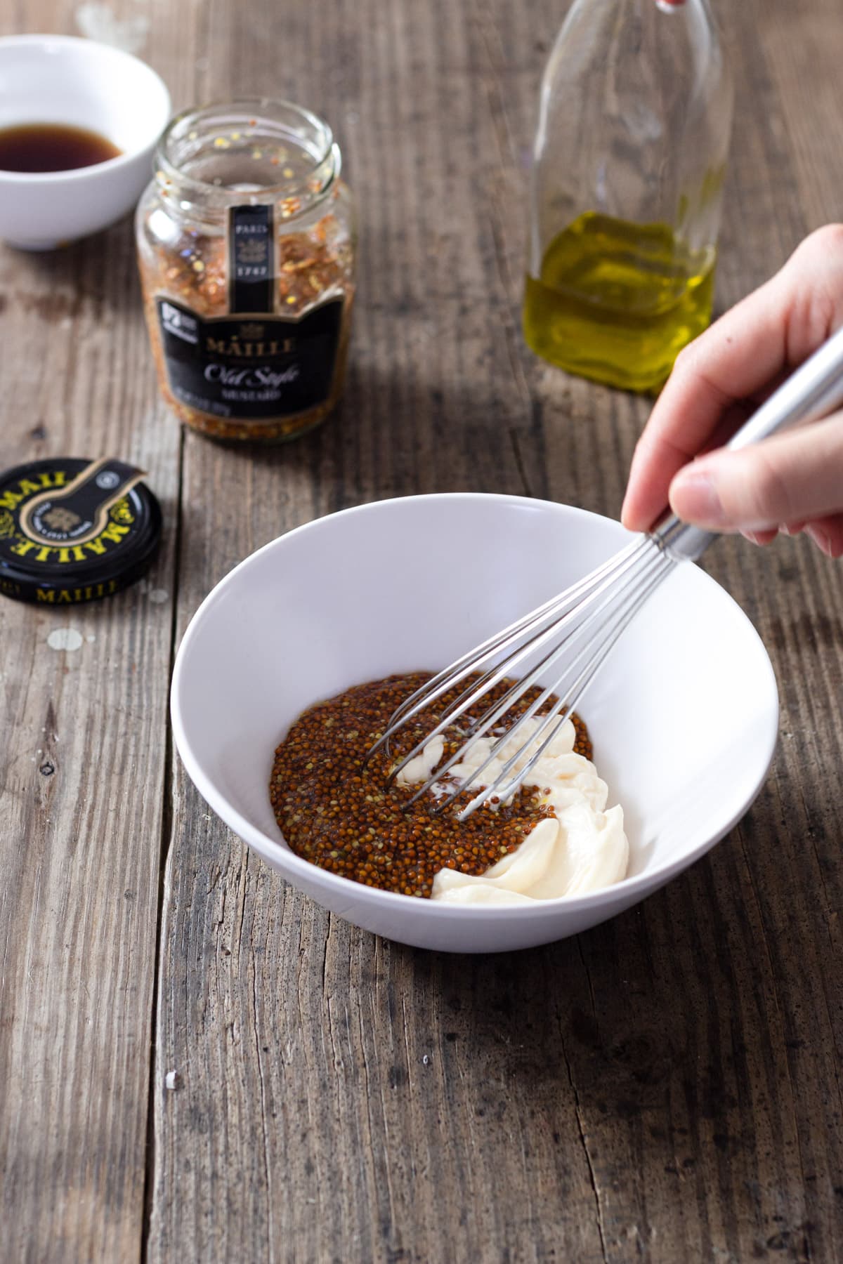 Whisk in a bowl of mayo, mustard seeds, oil and vinegar with ingredients in the background.