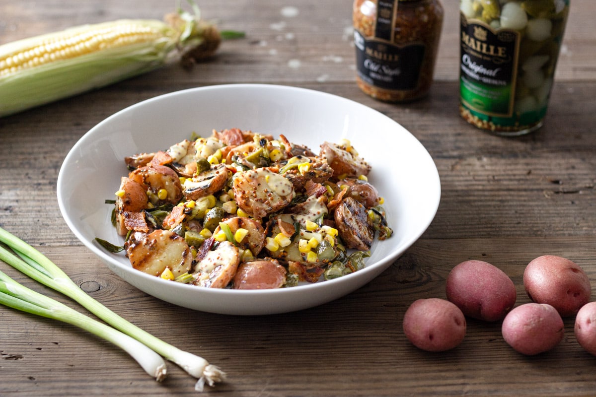 A bowl of Grilled Potato Salad with corn, bacon, and scallions on a wood surface surrounded by raw ingredients.