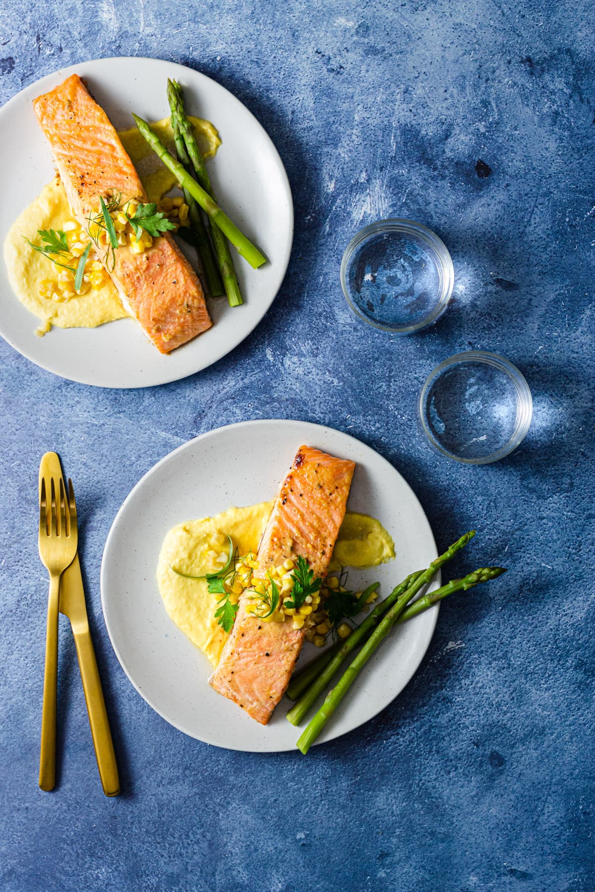 Two plates of Miso Butter Salmon over Corn Puree next to asparagus on light grey plates with a vibrant blue background next to water glasses and utensils.