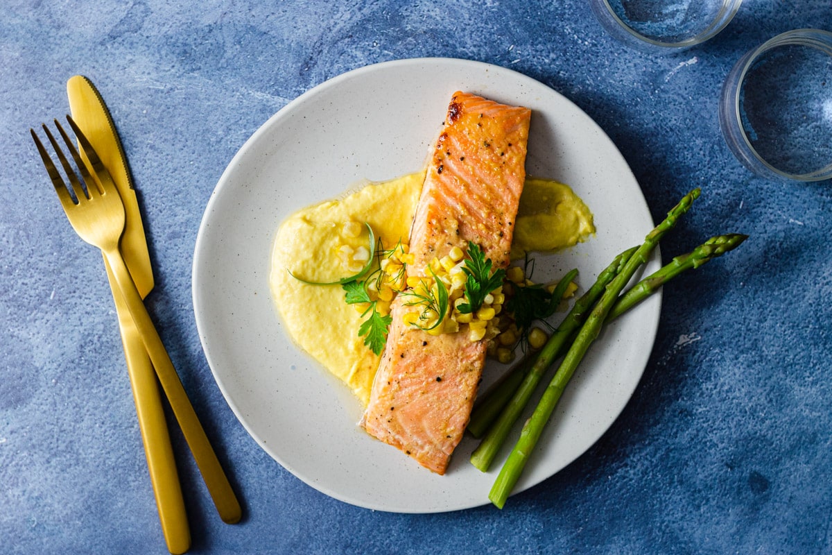 Overhead view of a plate of salmon over a corn puree next to asparagus surrounded by a fork and knife and water glasses.