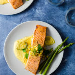 Miso Butter Salmon over Corn Puree next to asparagus on a light grey plate with a vibrant blue background.