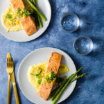 Two plates of Miso Butter Salmon over Corn Puree next to asparagus on light grey plates with a vibrant blue background next to water glasses and utensils.