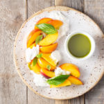 Overhead view of a plate of peach and burrata salad with fresh basil leaves and a cup of basil oil.