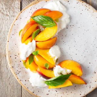 Overhead view of a rustic plate topped with sliced peaches, chunks of burrata cheese and fresh basil leaves with a cup of basil oil in the corner.