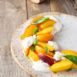 Angeled view of a rustic plate topped with sliced peaches, mounds of burrata cheese and fresh basil leaves.