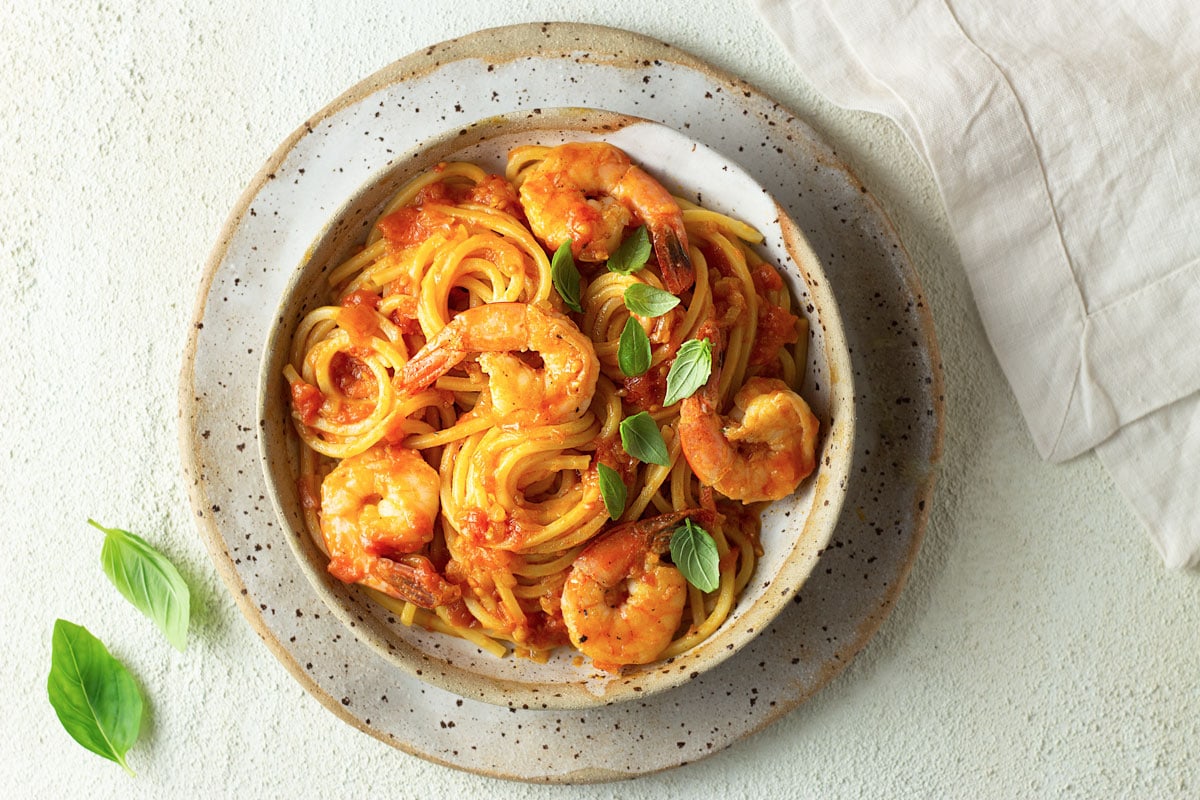 Overhead view of a bowl of spaghetti, shrimp and tomatoes on a rustic, cream colored surface.