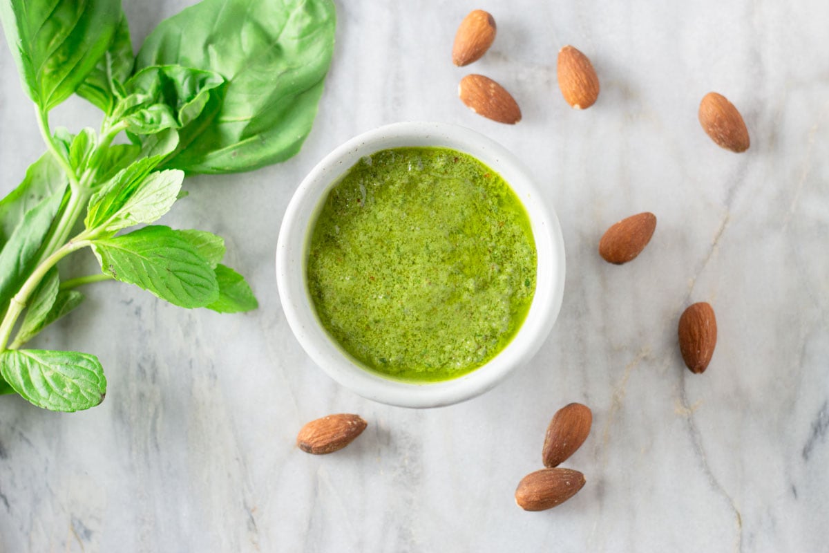 Overhead view of a bowl of pesto on a marble surface surrounded by almonds, basil and mint leaves.