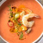 Overhead view of a bowl of tomato gazpacho topped with shrimp, sliced tomatoes and chives on a wood surface.