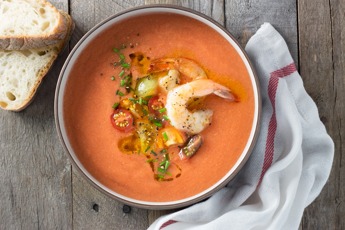 Overhead view of a bowl of tomato gazpacho topped with shrimp, sliced tomatoes and chives surrounded by slices of bread and a dish towel.