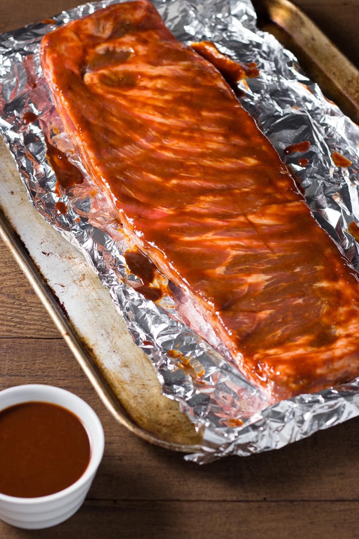 Rack of raw pork spare ribs on foil on a sheet tray brushed with barbecue sauce.