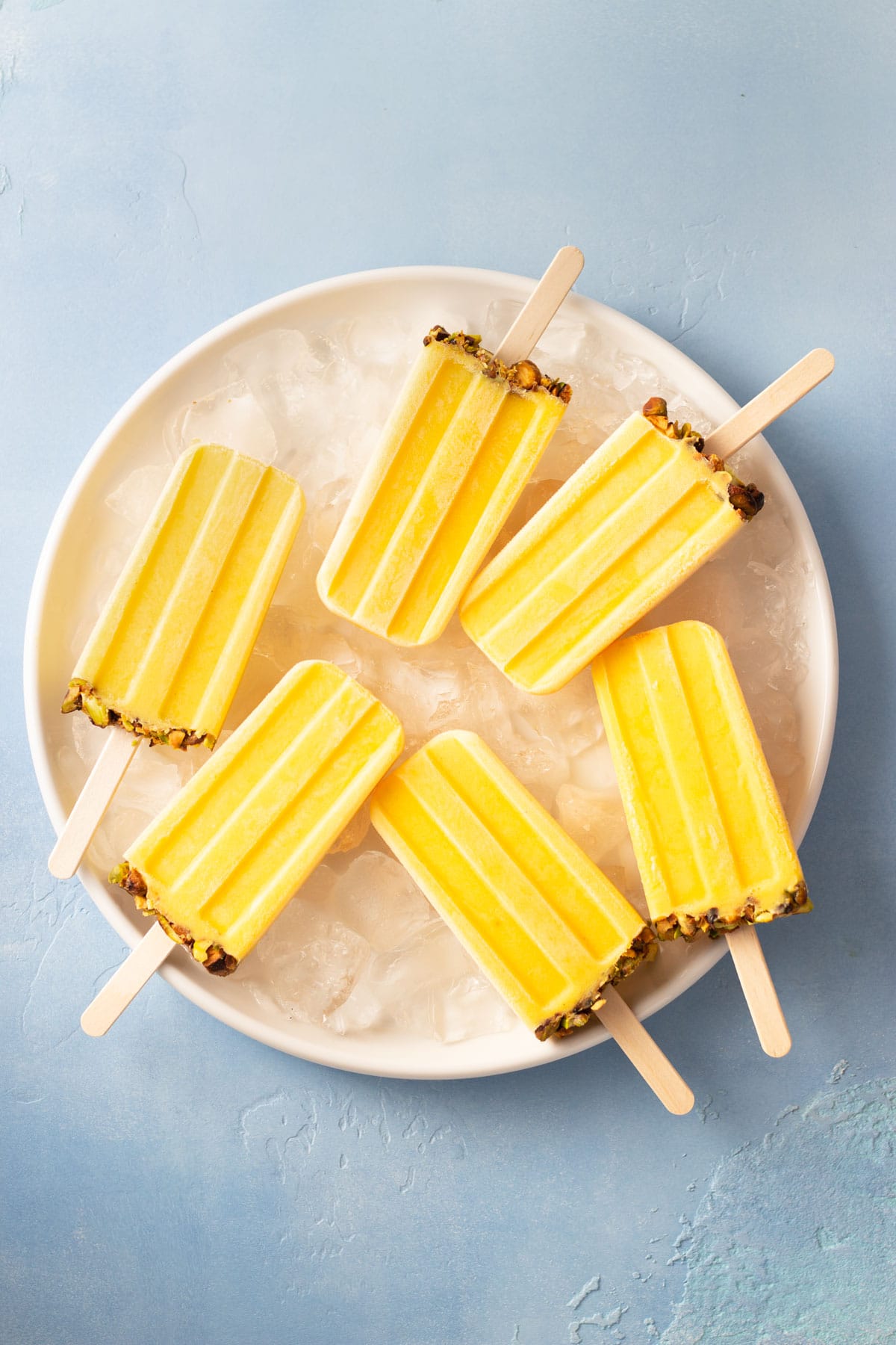 Overhead view of a group of Mango Lassi Popsicles on a plate of ice on a blue surface.