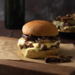 Straight on view of a Mushroom Swiss Burger with grilled onions on a cutting board on a dark wood surface with a burger and a bottle in the background, and a dark grey textured backdrop.