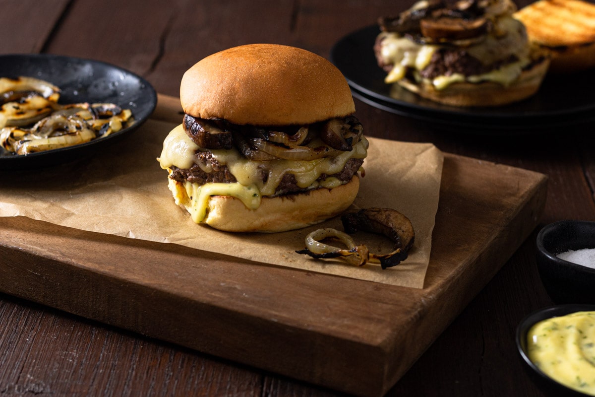 A burger topped with cheese, grilled onions and mushrooms on parchment on a cutting board next to a bowl of grilled onions and may with another burger in the background.