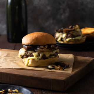 Straight on view of a Mushroom Swiss Burger with grilled onions on a cutting board on a dark wood surface with a burger and a bottle in the background, and a dark grey textured backdrop.