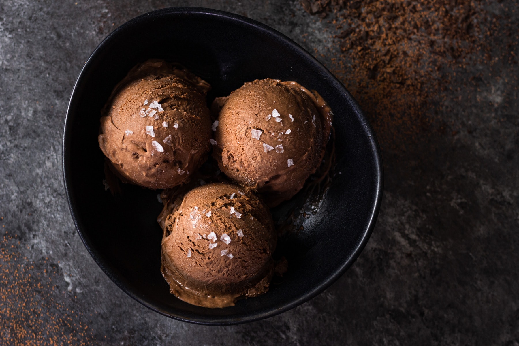 Overhead view of three scoops of chocolate olive oil ice cream topped with salt in a black bowl on a dark grey surface.