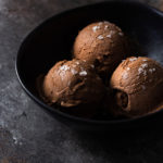 Black bowl of scoops of chocolate olive oil ice cream topped with salt on a dark grey surface.