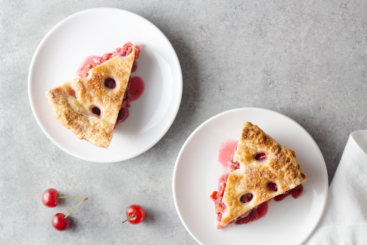 Overhead view of two slices of cherry pie on white dessert plates on a light grey surface.