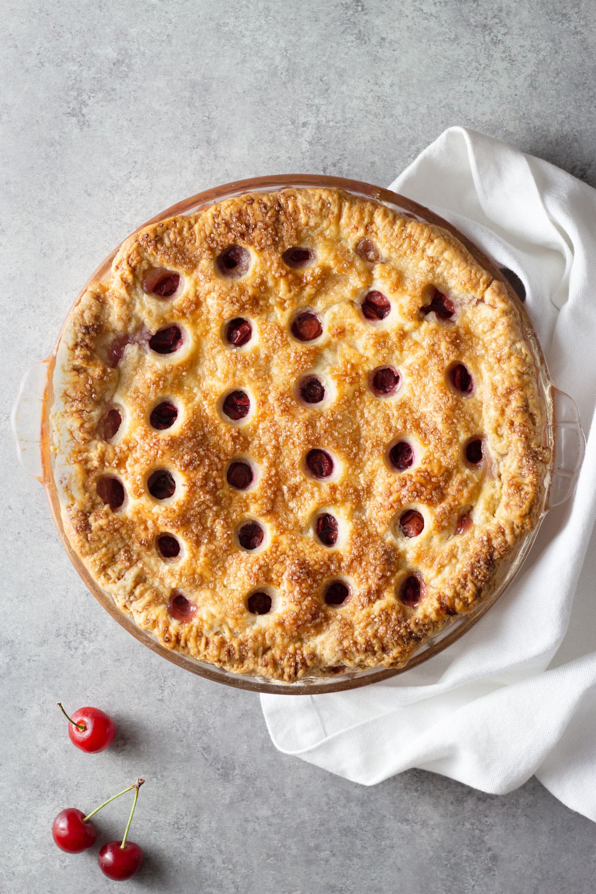Overhead view of a Sour Cherry Pie with circles cut out of the top crust on a grey surface next to cherries and a dish towel.
