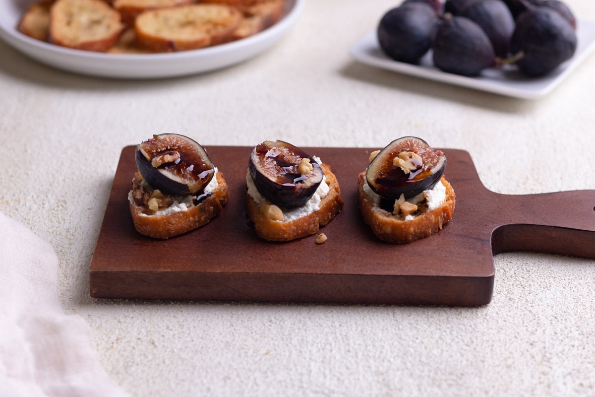 Fig and goat cheese crostini with balsamic on a wood cutting board on a cream, textured surface.