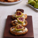 Fig and goat cheese crostini with walnuts and honey on a small wood cutting board on a cream, textured surface.