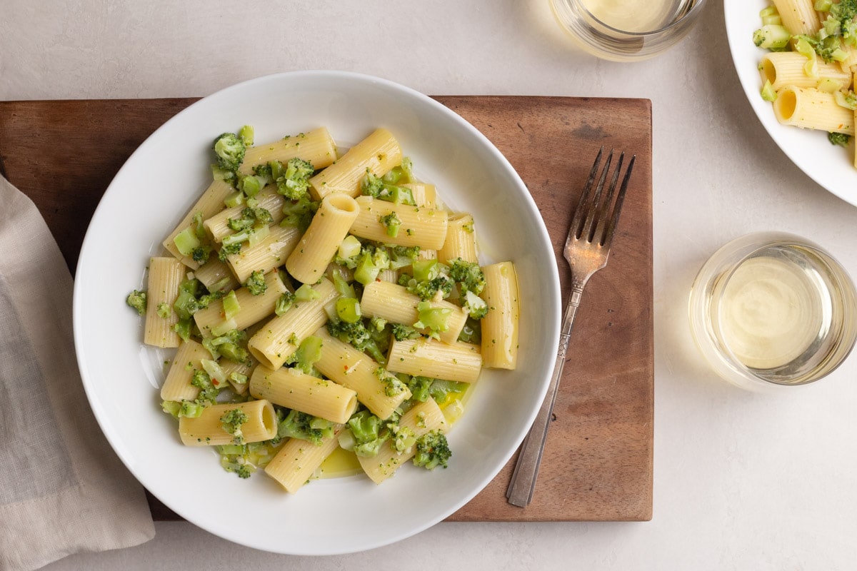 Italian Broccoli Pasta in a white bowl on a wood board on a cream surface surrounded by a dishcloth, white wine, and a fork.