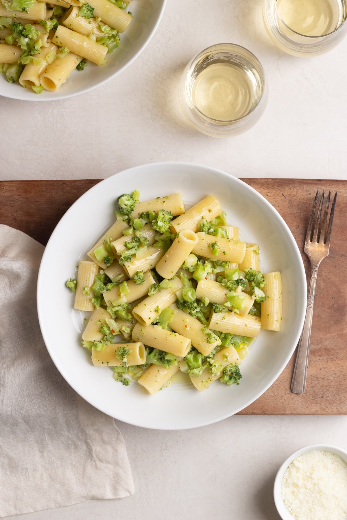 Italian Broccoli Pasta in a white bowl on a wood board on a cream surface surrounded by a dishcloth, white wine, a fork and grated cheese.