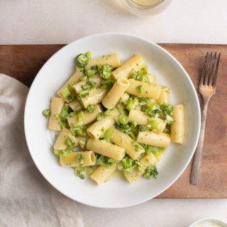 Broccoli Pasta in a white bowl on a wood board on a cream surface surrounded by a dishcloth, white wine, a fork and grated cheese.