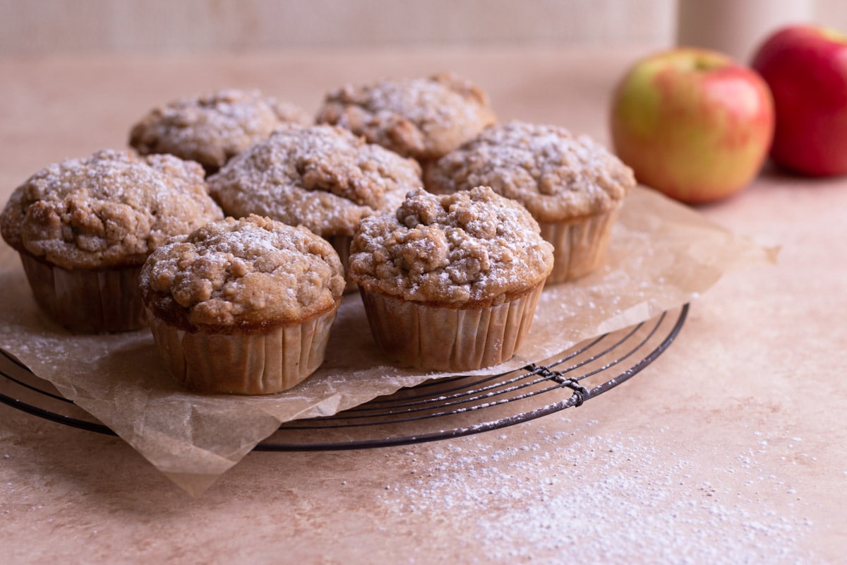 Spiced Apple Sour Cream Muffins with Crumb Topping on a cooling rack with apples in the background.