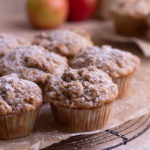A group of apple crumb muffins on brown parchment over a round cooling rack with apples in the background.
