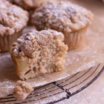 Close up view of a cut open apple crumb muffin with more muffins in background on a cooling rack.