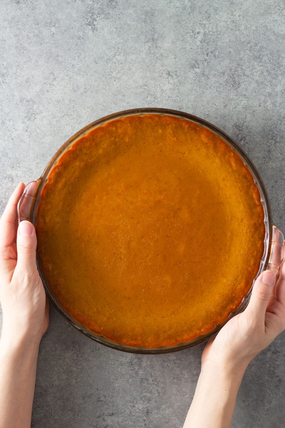 Overhead view of hands holding a pumpkin pie on a grey surface.