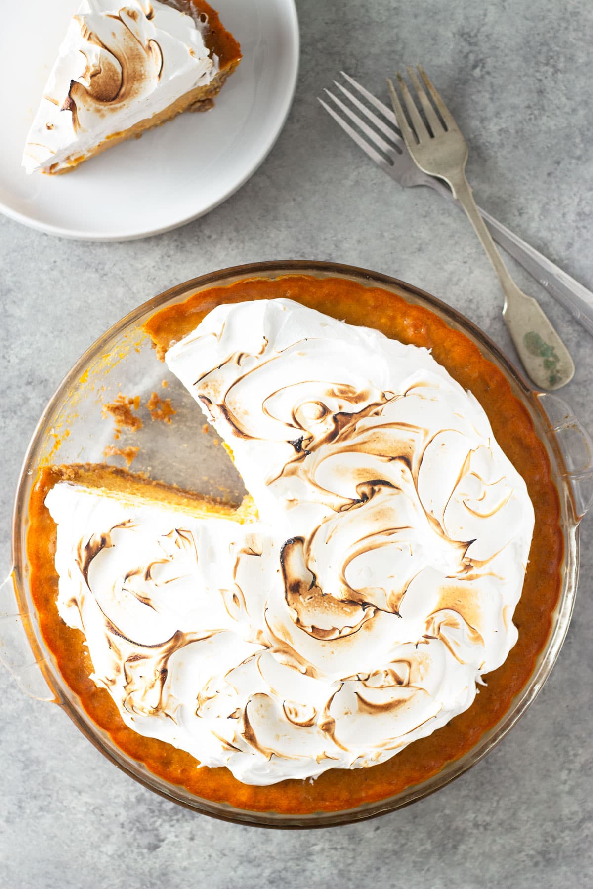 Overhead view of a pumpkin meringue pie with a slice cut out on a plate to the side on a grey, textured surface.
