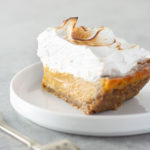 Straight on view of a slice pumpkin meringue pie next to a fork on a grey, textured surface.