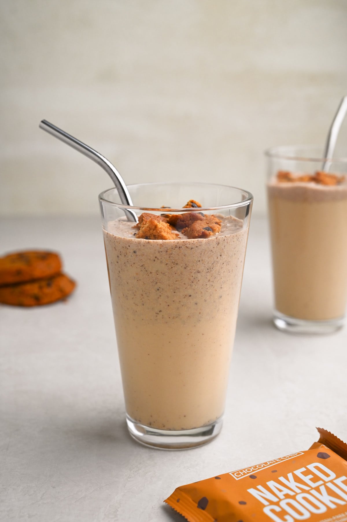 Straight on view of two glasses of a cookie protein shake with metal straws, cookies on top and cookies in the background.