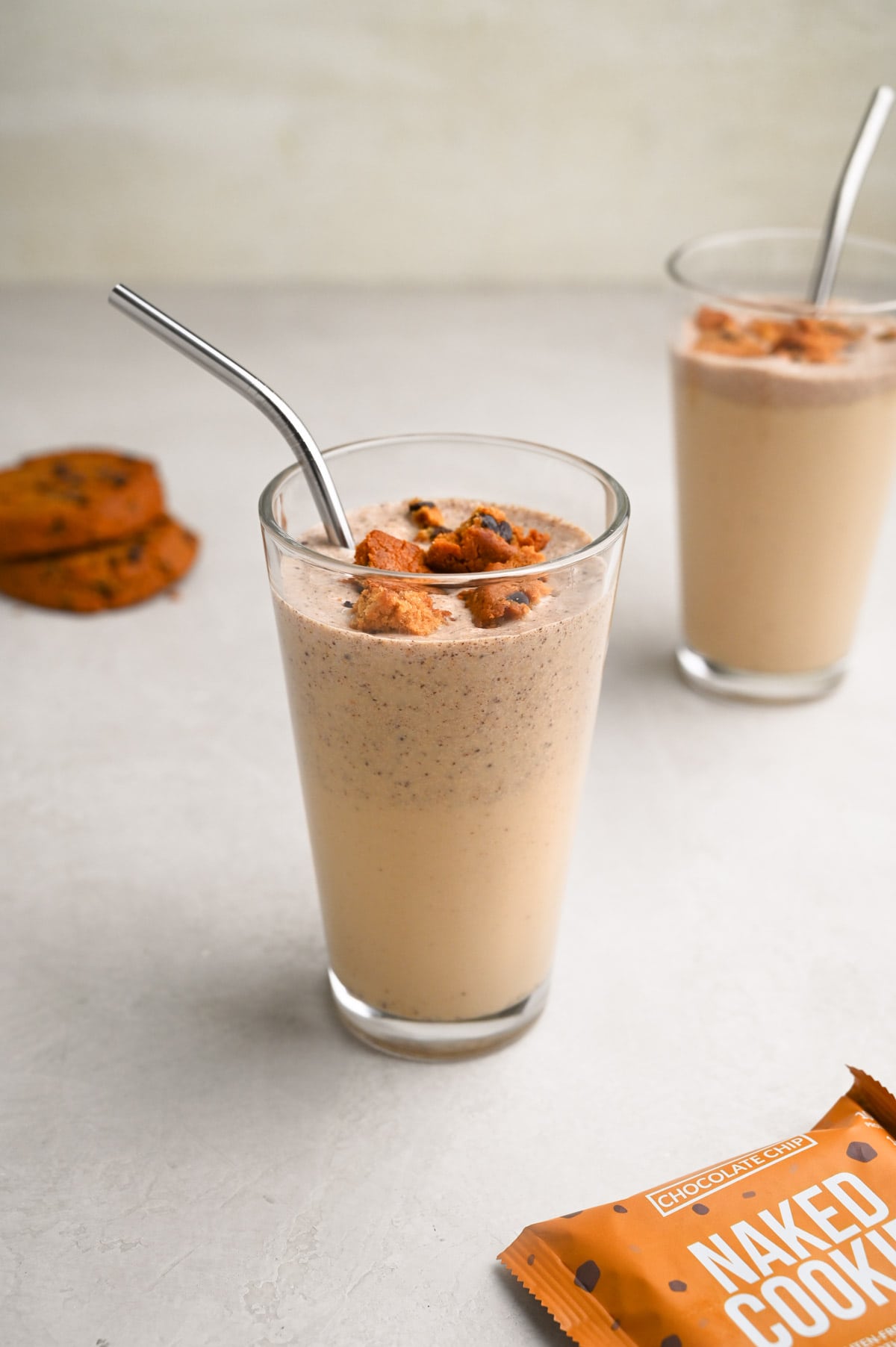 Angled view of two glasses of cookie shakes with protein chocolate chip cookies crumbled on top and whole cookies in the background.